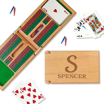 Personalized cribbage game board engraved with an initial above a name on a white background, Next to it is an opened game revealing cribbage track, pegs and a deck of cards. 